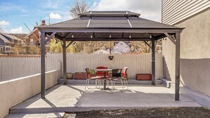Un patio cubierto con una mesa y sillas, una cerca blanca de piquete y una casa al fondo.