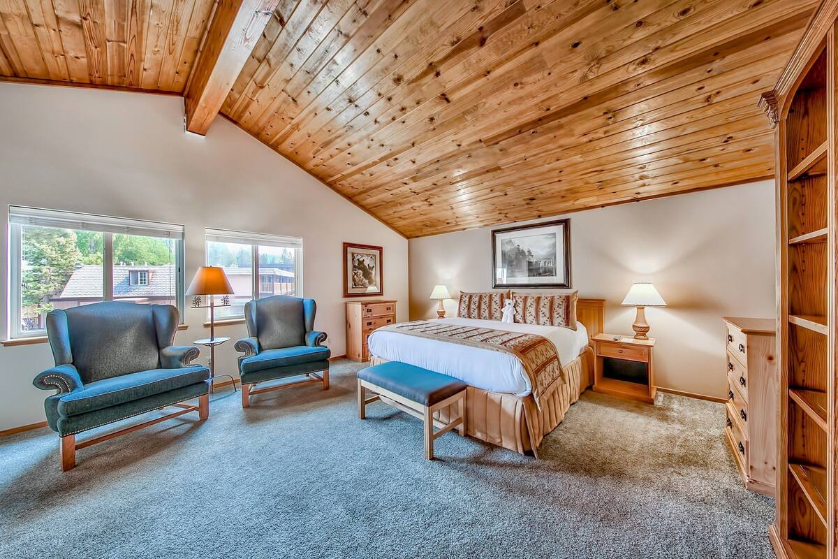 Un dormitorio con techo de madera, dos sillones, una cama con colcha blanca y marrón, una mesita de noche con lámpara y un aparador.