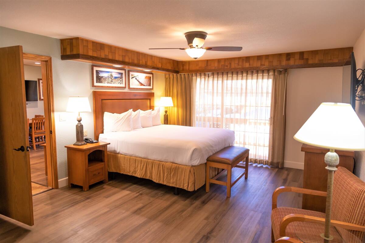 Habitación de hotel con una cama grande, cabecera de madera, dos mesitas de noche con lámparas, un banco de madera, un escritorio con lámpara, un ventilador de techo y una ventana con cortinas.