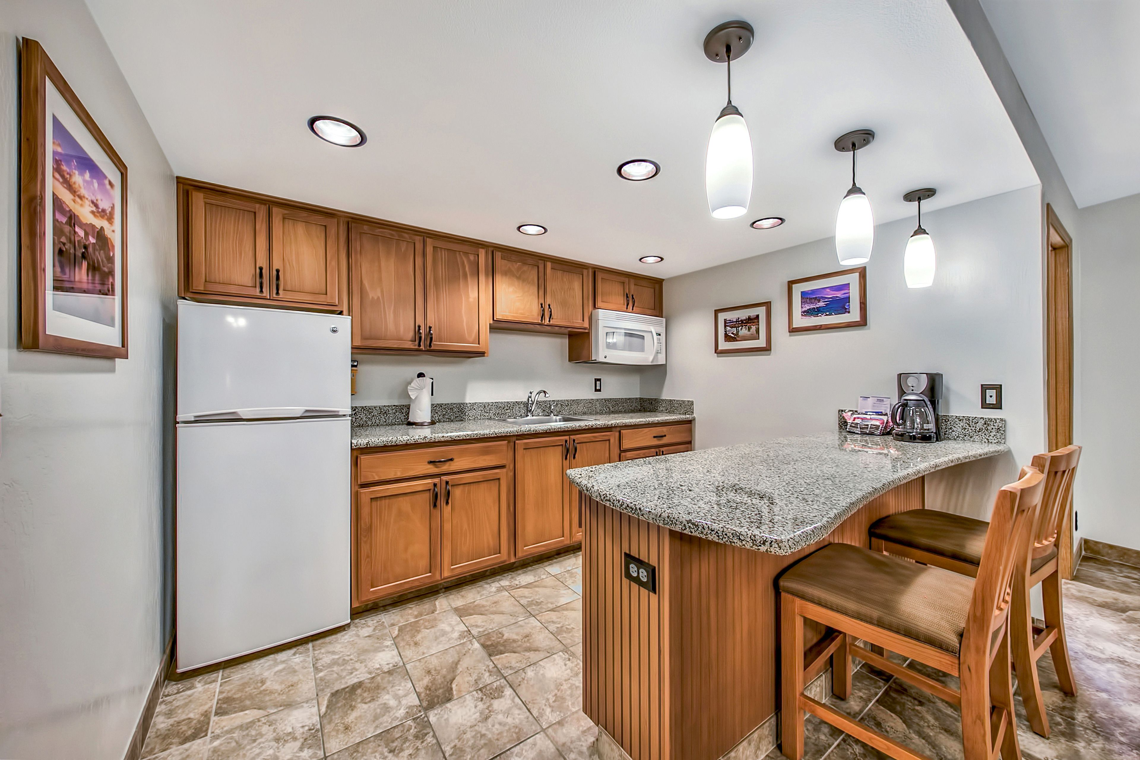 A kitchen with wooden cabinets, a white refrigerator, a microwave, granite countertops, and a bar area with two stools.