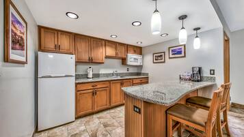A kitchen with wooden cabinets, a white refrigerator, a microwave, granite countertops, and a bar area with two stools.