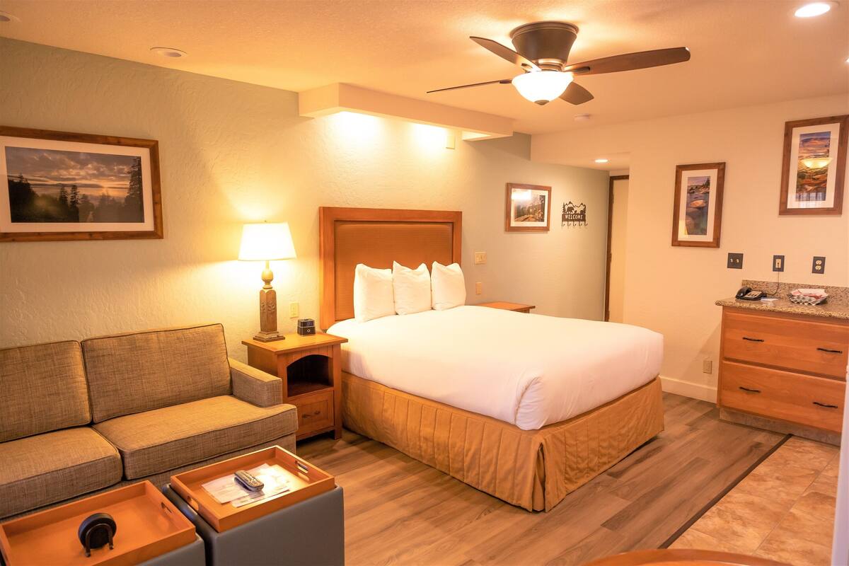 Habitación de hotel con cama, sofá, escritorio con lámpara, ventilador de techo e imágenes enmarcadas en las paredes.