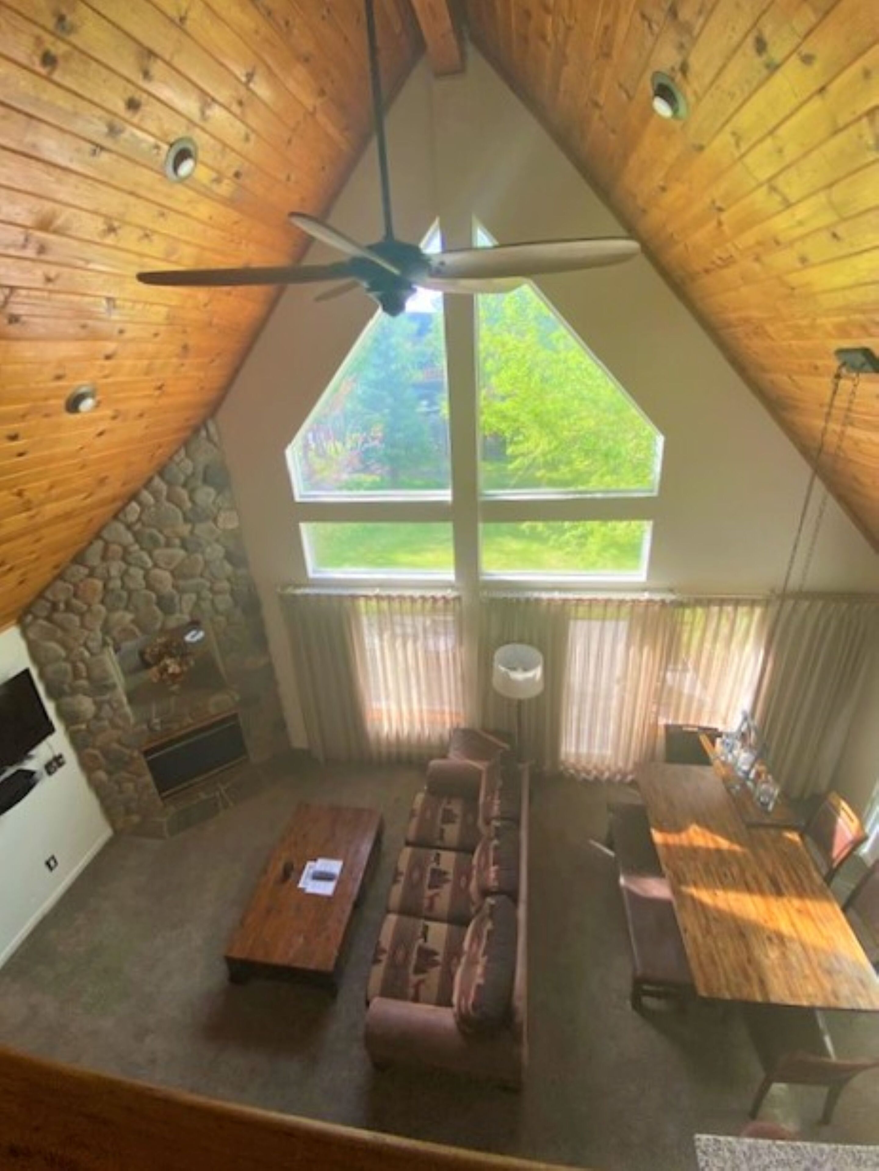 A cozy attic room with a sloped ceiling, a stone fireplace, a sofa, a dining table, and a large window offering a view of greenery.