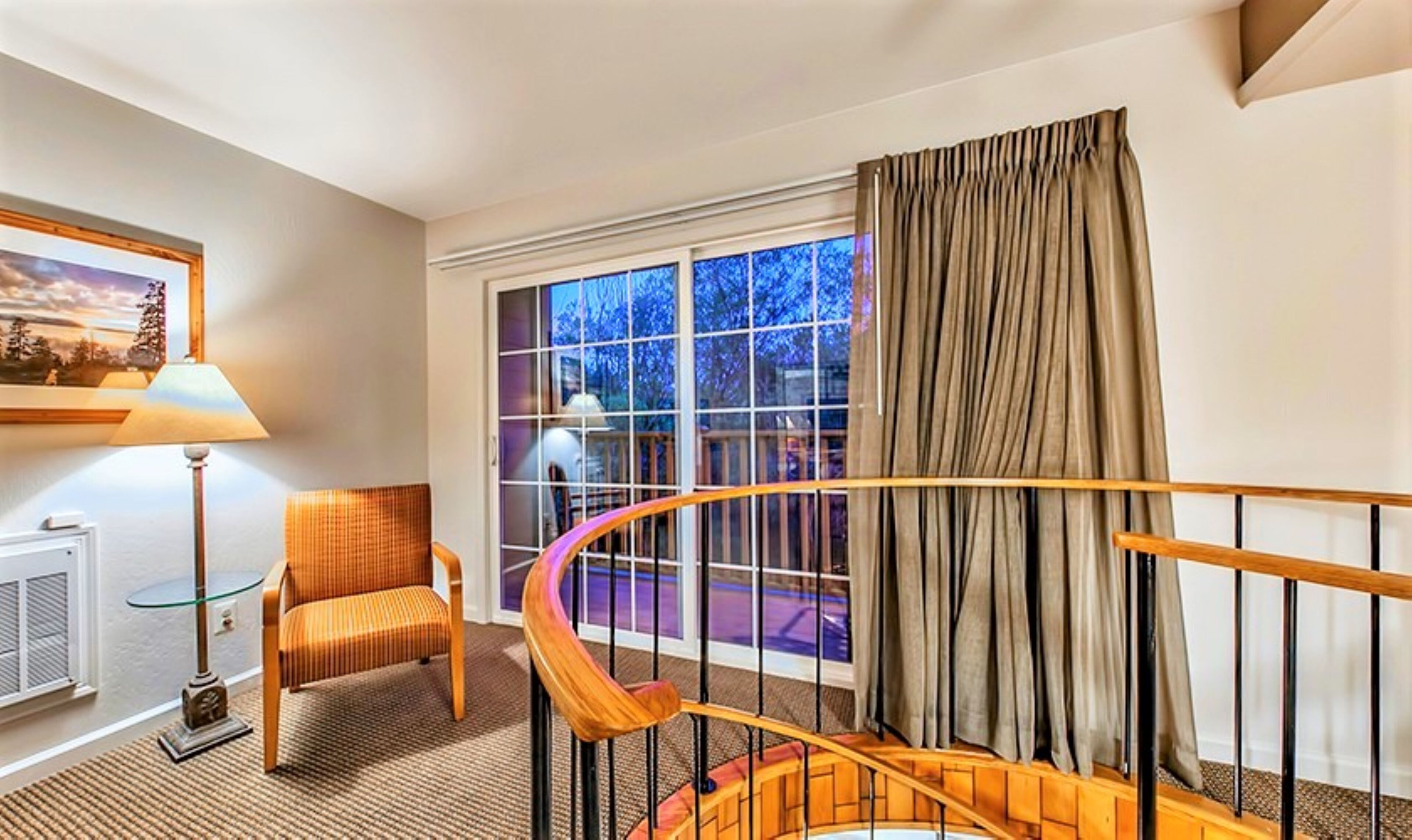 A hotel room with a view of trees through large windows, a wooden staircase, a chair, a lamp, and a radiator.