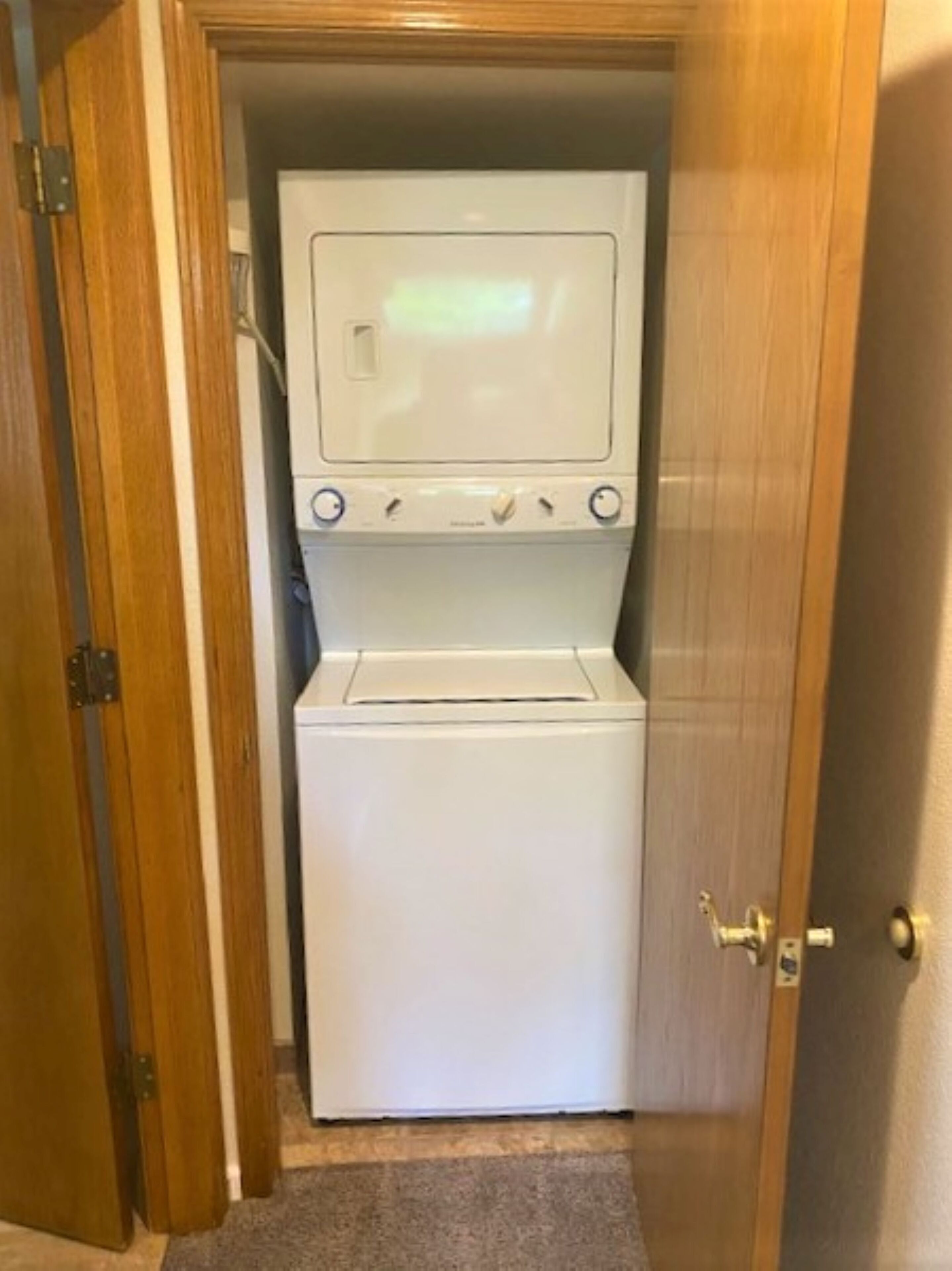 Une petite machine à laver blanche, placée dans un espace réduit et fermé, avec une porte en bois et un sol recouvert de moquette.