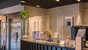 A reception desk with a computer, a vase of flowers, a digital thermometer, and a sign. - Atlas Hotel Brussels (Brussels)