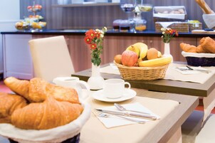 Un desayuno con cruasanes, una cesta de frutas y un florero con flores sobre una mesa.