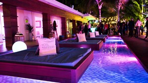 Een hotel lobby met een geruite vloer, meerdere bedden met kussens waarop 'HOTEL MARSEILLE' staat, en een zwembadgedeelte met lichtslingers.