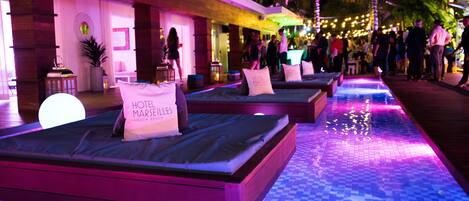 A hotel lobby with a checkered floor, multiple beds with pillows labeled "HOTEL MARSEILLE," and a pool area with string lights.