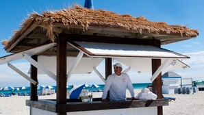 Een strandtentje met rieten dak, een persoon erachter en strandparasols op de achtergrond.