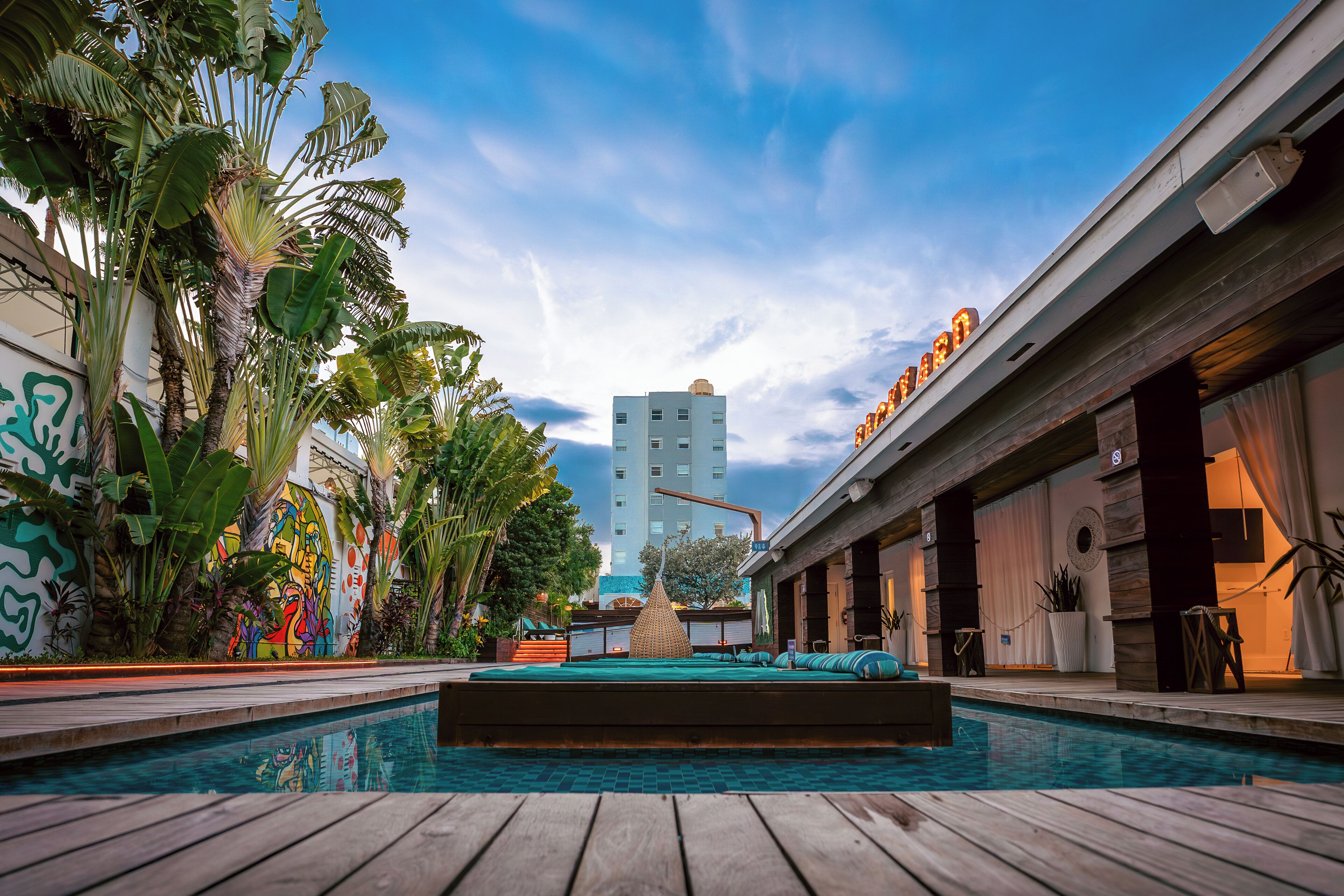 A pool area with a wooden deck, surrounded by lush greenery and a colorful mural. A tall building is visible in the background.