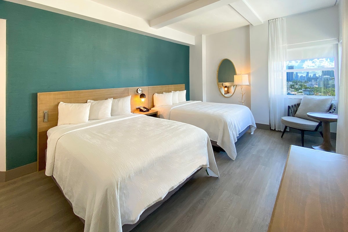 A hotel room with two beds, a sitting area, a view of the city, and a large mirror.