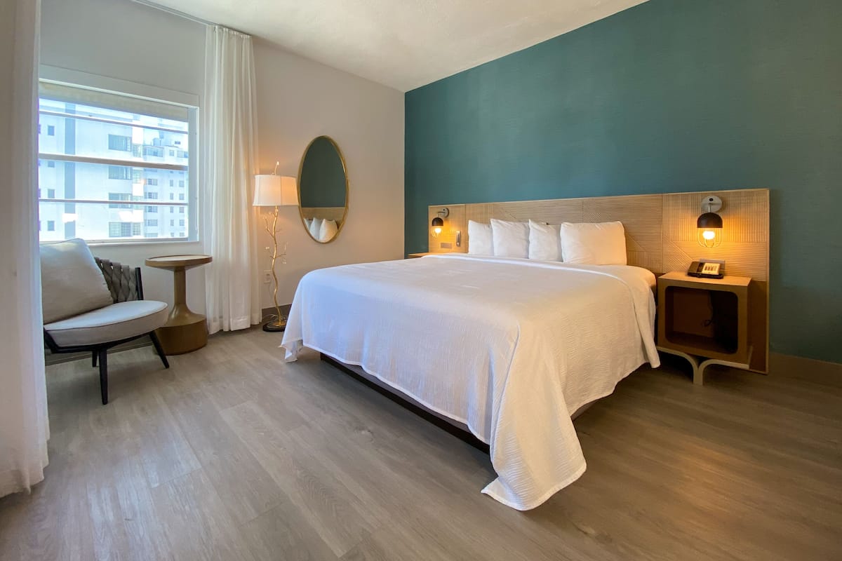 A hotel room with a large bed, a chair, a small table, a lamp, a mirror, and a nightstand with a phone.
