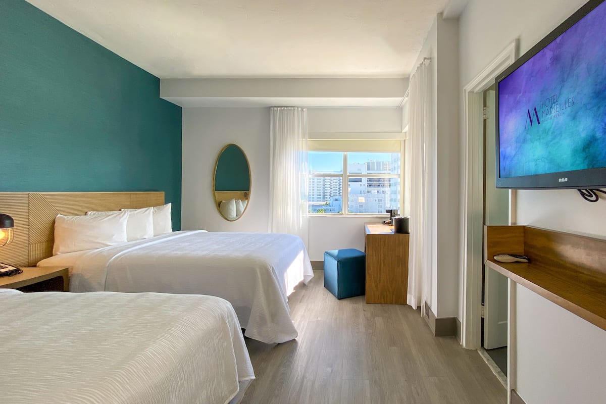 A hotel room with two beds, a TV, a desk, and a view of the city.