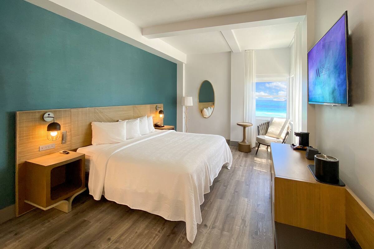 A hotel room with a large bed, a desk, a chair, a TV, and a view of the ocean.