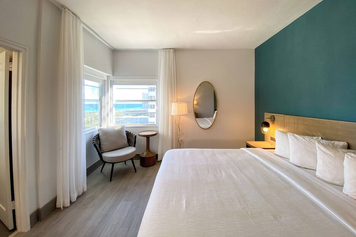 A hotel room with a large bed, a chair, a small table, a lamp, a mirror, and a view of the ocean.