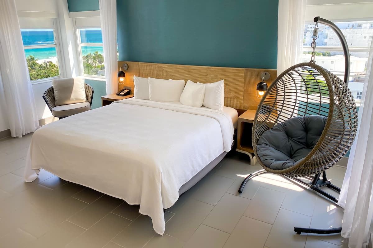 A hotel room with a large bed, a hanging chair, a nightstand, and a view of the ocean.