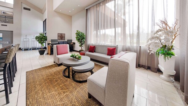A modern living room with a sofa, armchairs, a coffee table, and a dining area with chairs. There are potted plants and a large window with curtains. - Best Western Plus Redondo Beach Inn (Redondo Beach)
