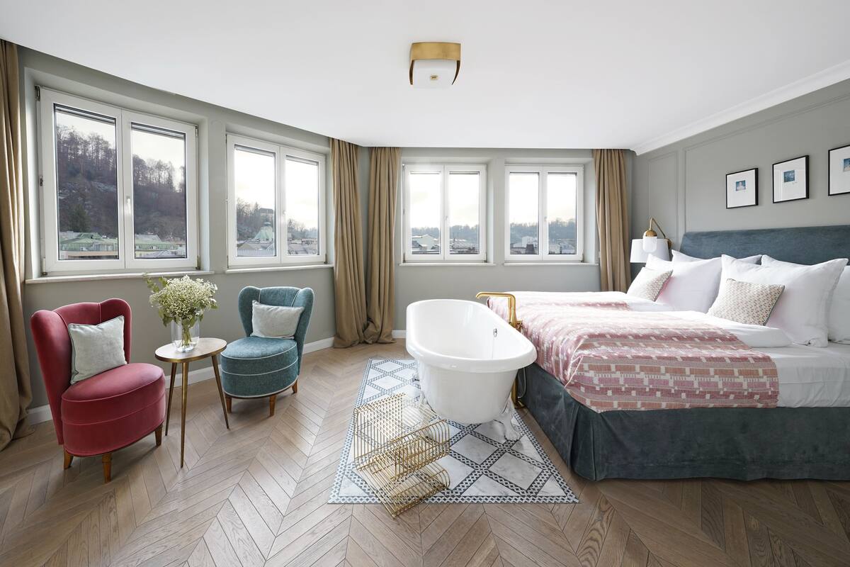 Um quarto espaçoso com uma cama grande, um espaço para sentar com uma cadeira e uma mesa pequena, uma banheira e amplas janelas com vista para o exterior.