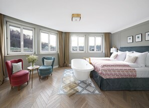 A spacious bedroom with a large bed, a sitting area with a chair and a small table, a bathtub, and large windows offering a view of the outdoors. - Numa Salzburg Mozart (Salzburg)