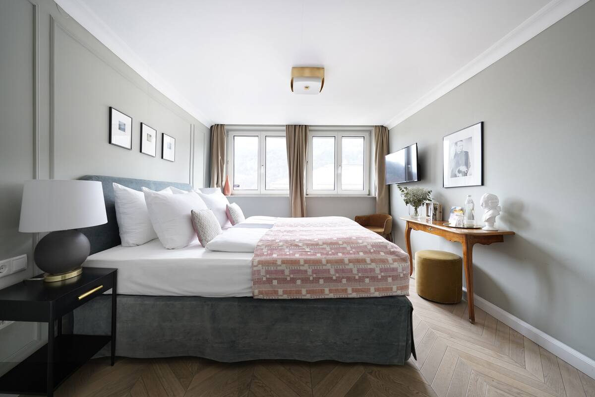 Quarto de hotel moderno com uma cama grande, uma mesinha de cabeceira com abajur, uma mesinha lateral de madeira e uma TV fixada na parede.