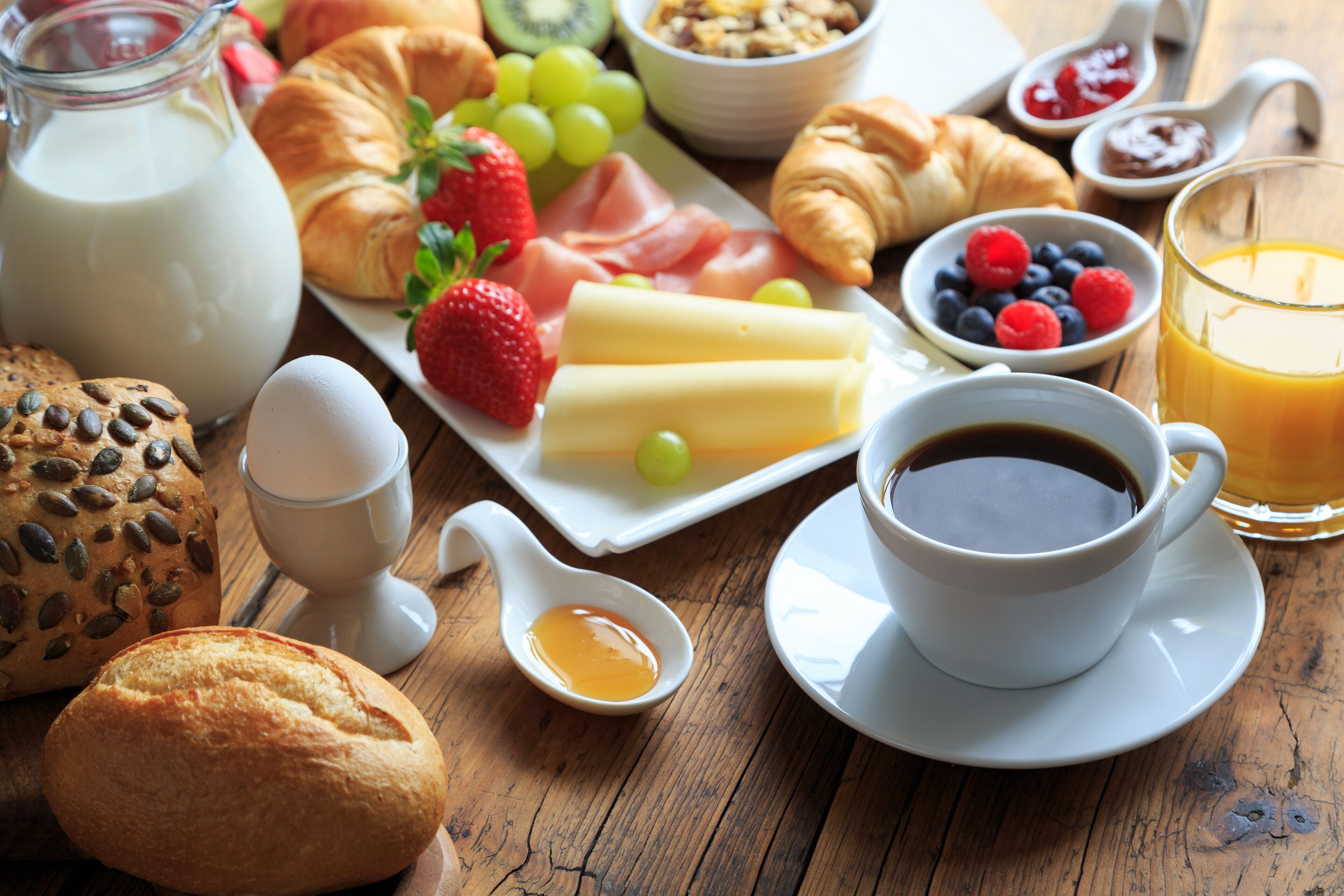 Desayuno con cruasanes, fresas, queso y una taza de café sobre una mesa de madera.