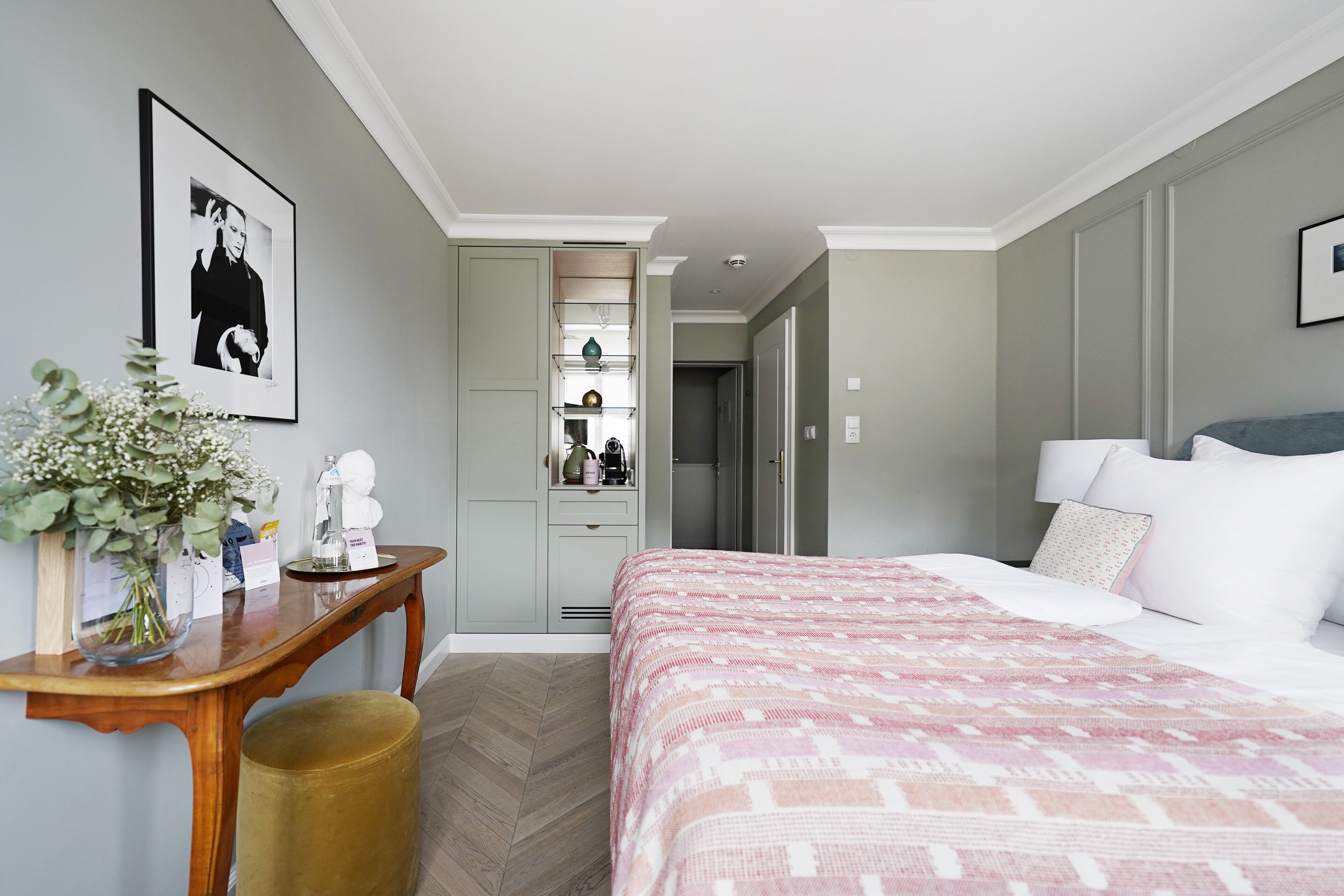 A hotel room with a bed, a desk with a vase of flowers, a framed picture on the wall, and a wardrobe.