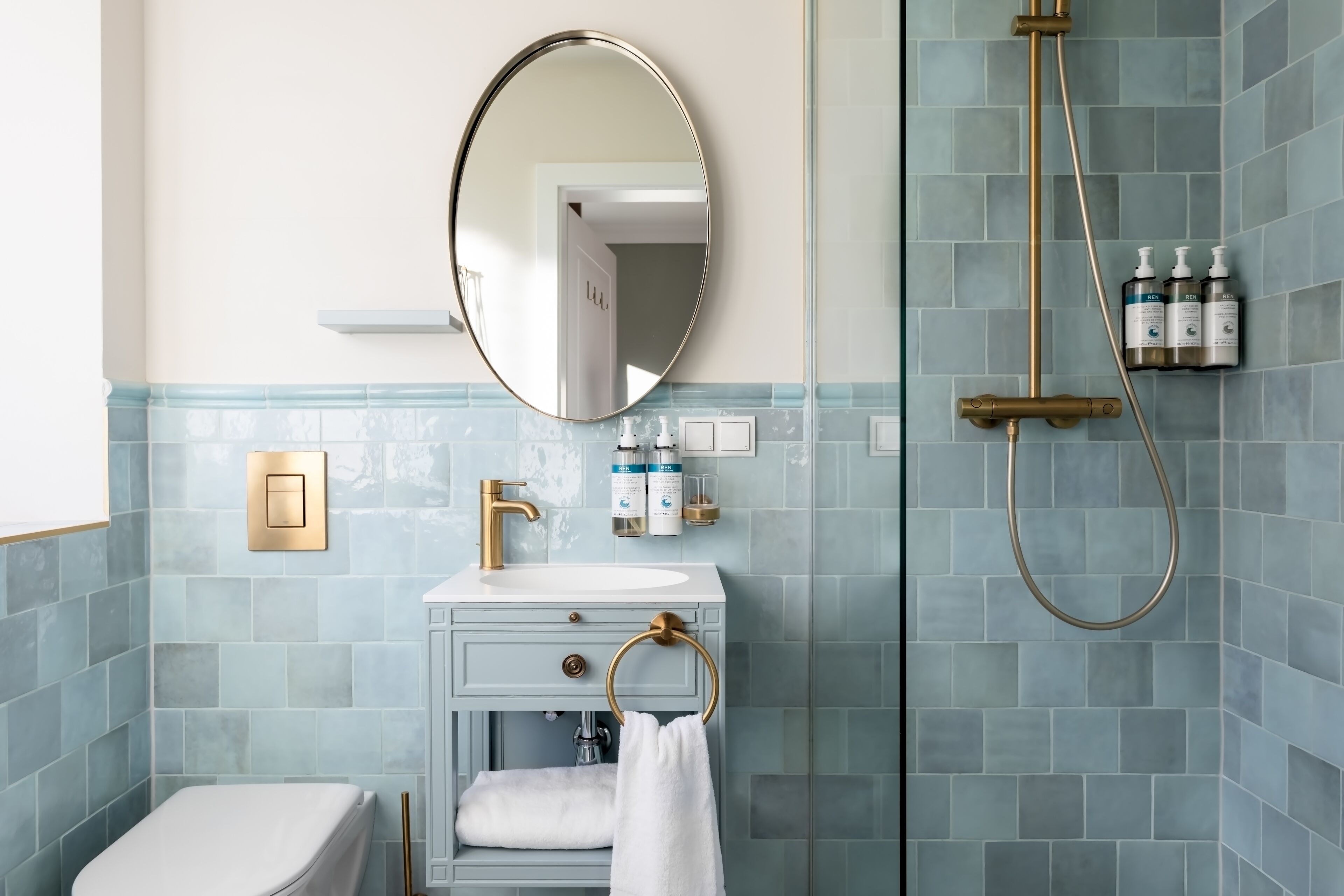 A bathroom with a white sink, gold faucet, and a mirror above it. There is a towel ring on the sink and a shower with a gold showerhead.