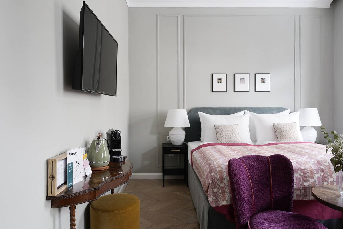 Quarto de hotel com uma cama, mesas de cabeceira, televisão e uma pequena mesa com abajur e objetos decorativos.