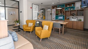 Eine moderne Hotelhalle mit einer Couch, zwei gelben Sesseln, einem Holztisch und einer Küchenzeile mit Regalen und einem Fernseher.