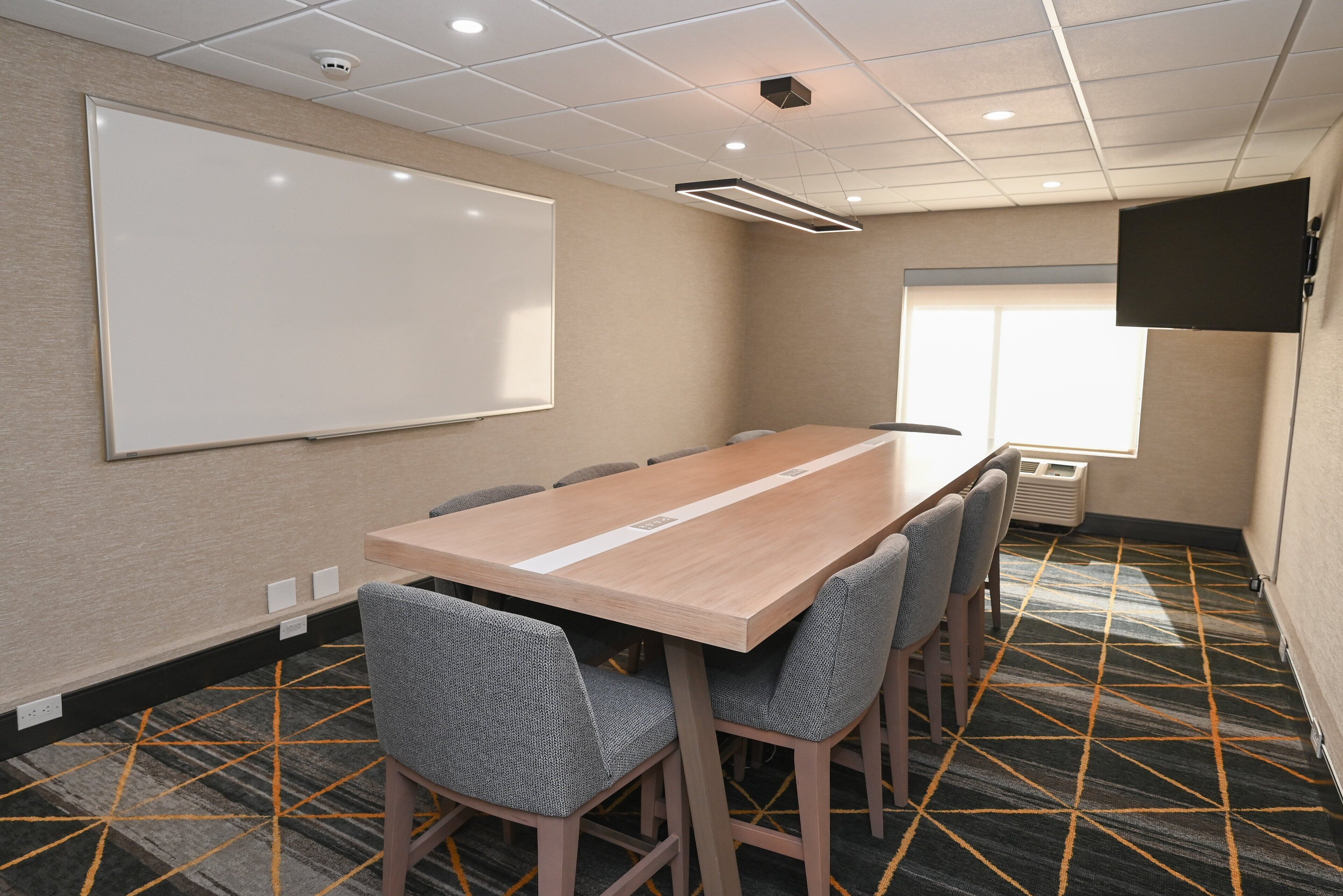 A conference room with a long table, six chairs, a whiteboard, a projector screen, and a patterned carpet.