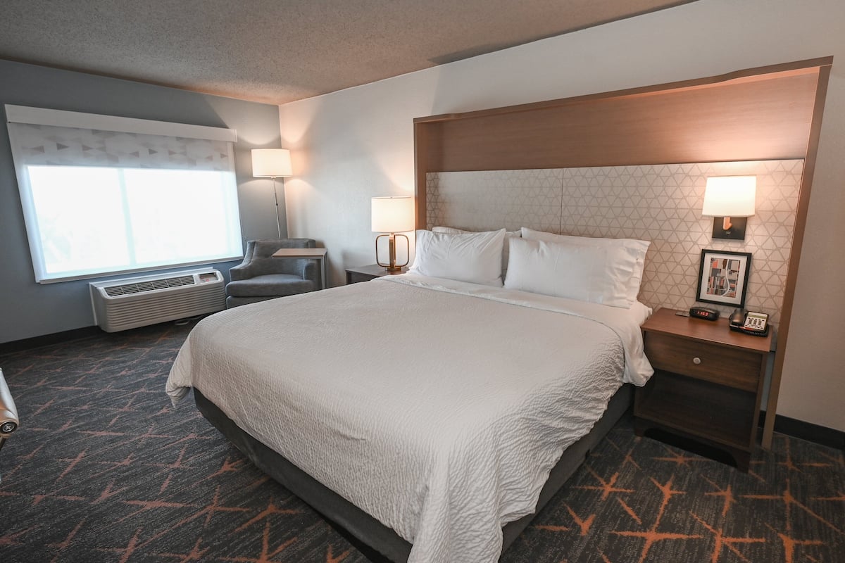 A hotel room with a large bed, two bedside tables with lamps, a chair, a window with blinds, and a wall-mounted air conditioner.