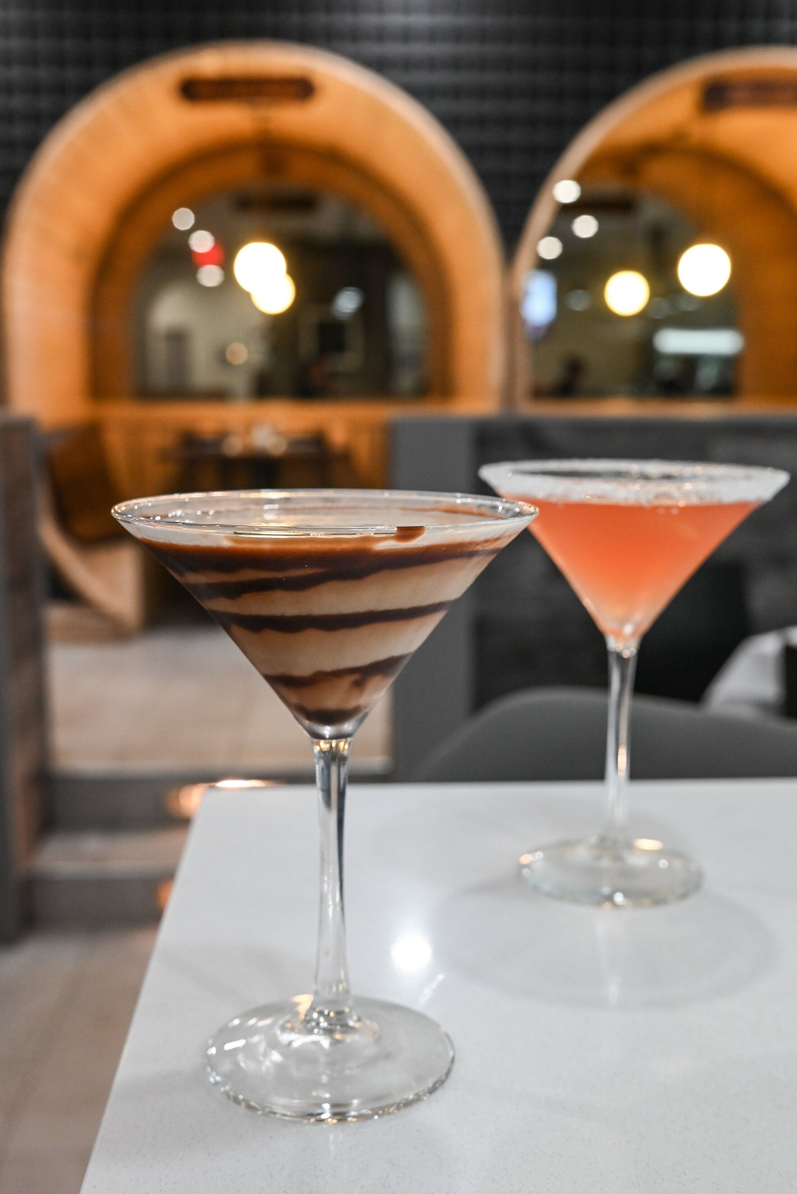Duas taças de martini sobre uma mesa branca, com um fundo desfocado mostrando estruturas de madeira arqueada.