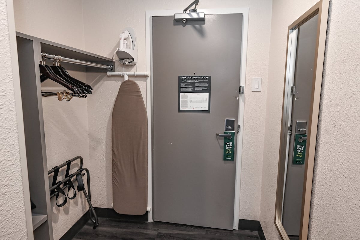 A hotel room with a gray door, an emergency evacuation plan sign, a coat rack with hangers, a towel rack, and a wheelchair.