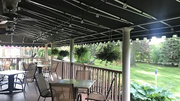 An outdoor patio with tables and chairs, a pergola with string lights, and a view of a well-maintained lawn and trees.