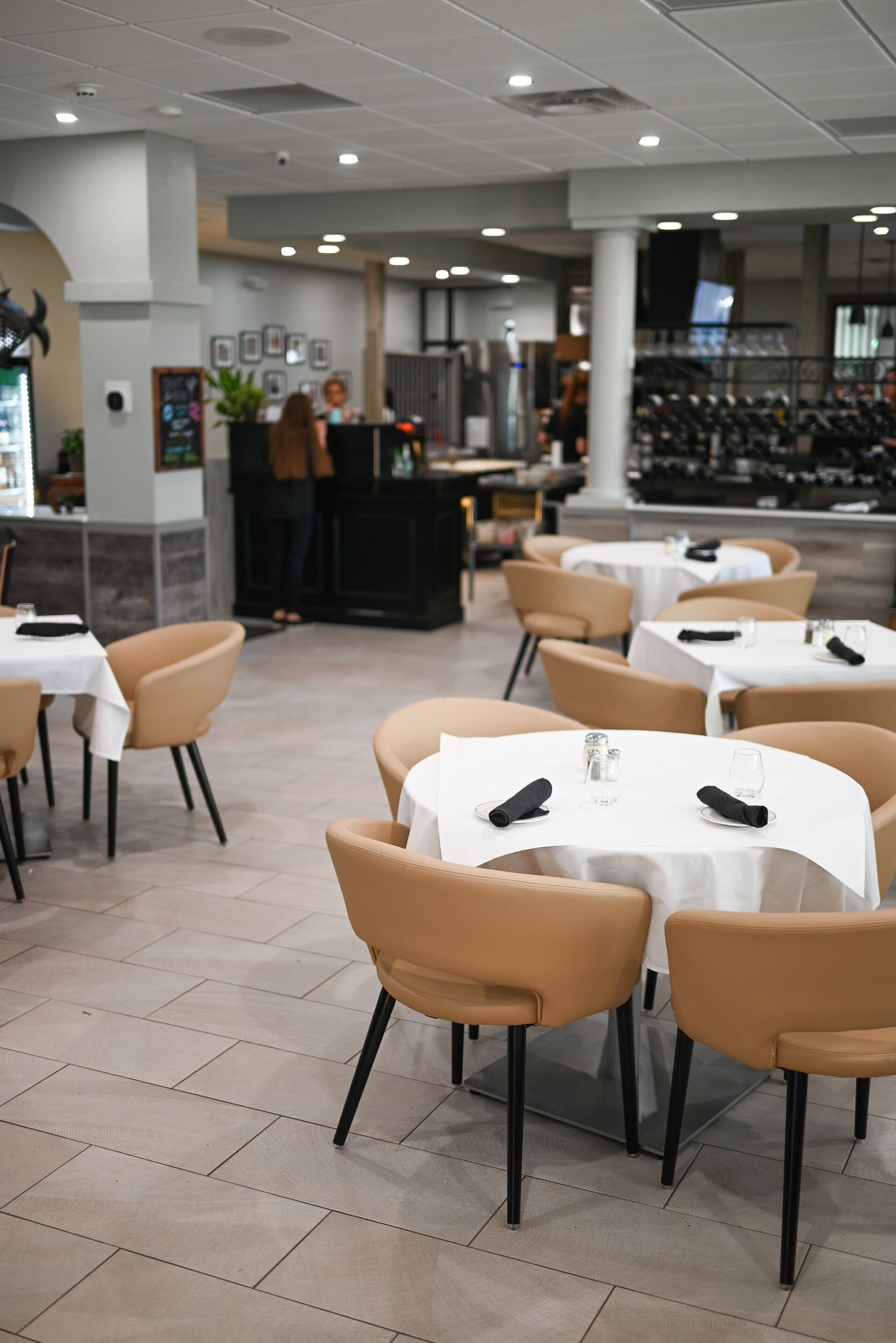L’intérieur d’un restaurant avec des tables rondes et des chaises de couleur beige, un bar à l’arrière-plan et un sol carrelé.