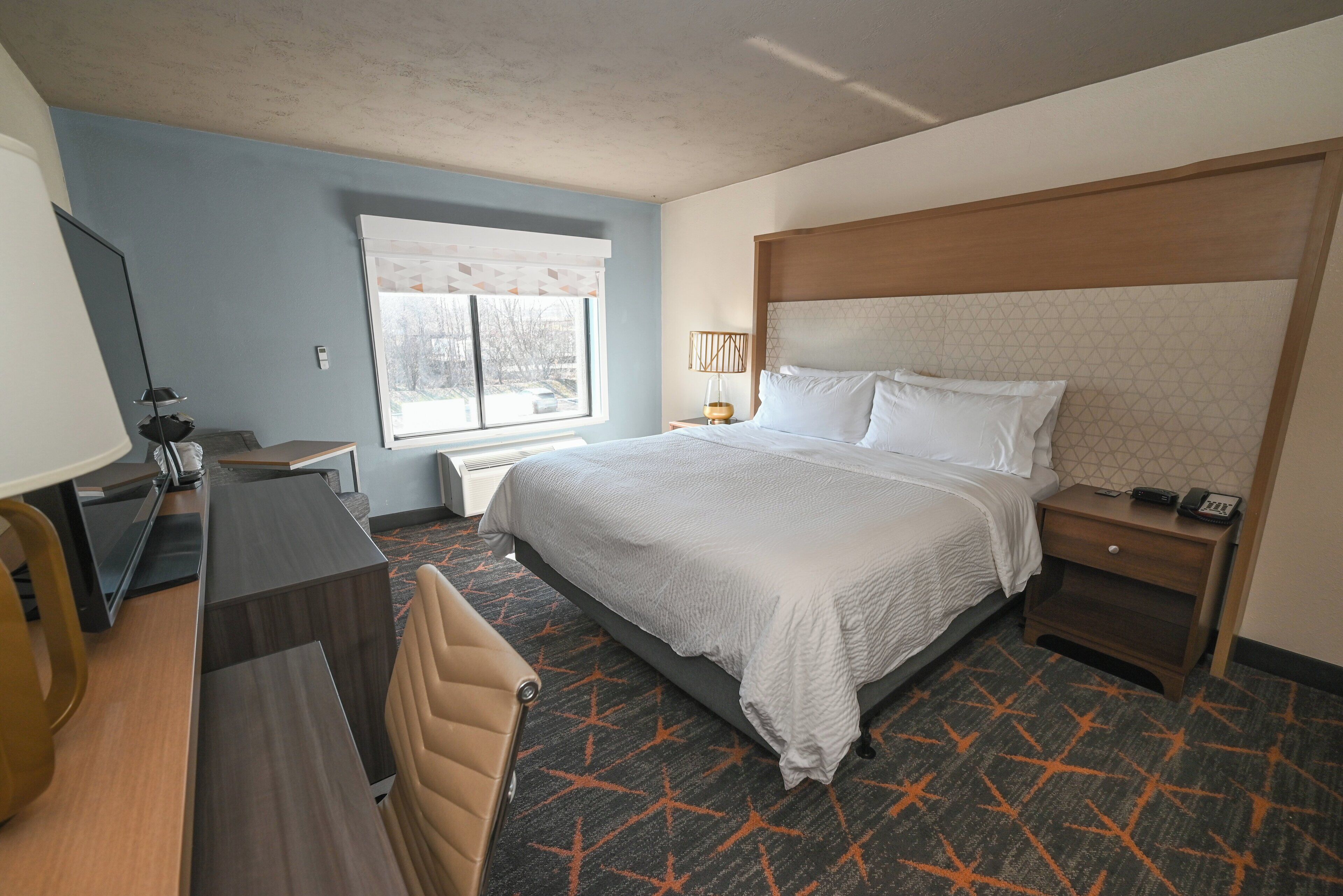 A hotel room with a bed, bedside table, lamp, desk, chair, and a window with a view.