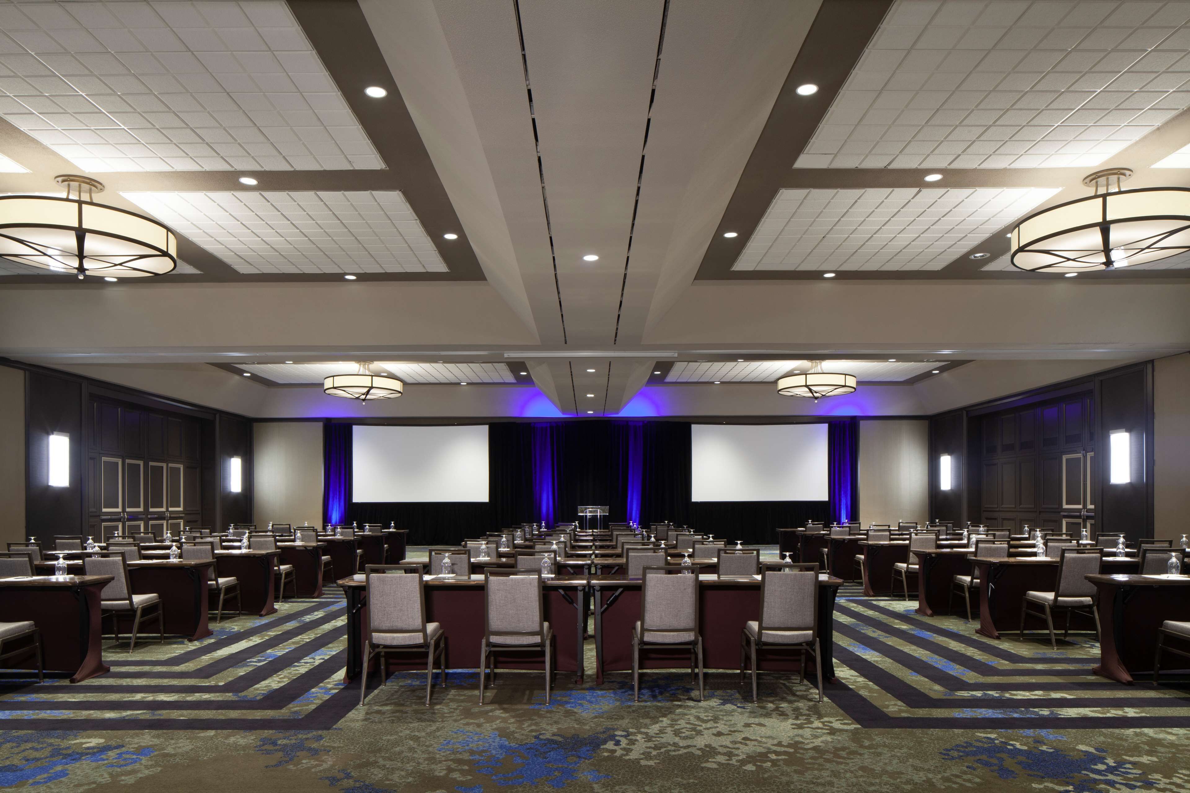 A conference room with rows of tables and chairs, a large screen, and a stage with curtains.