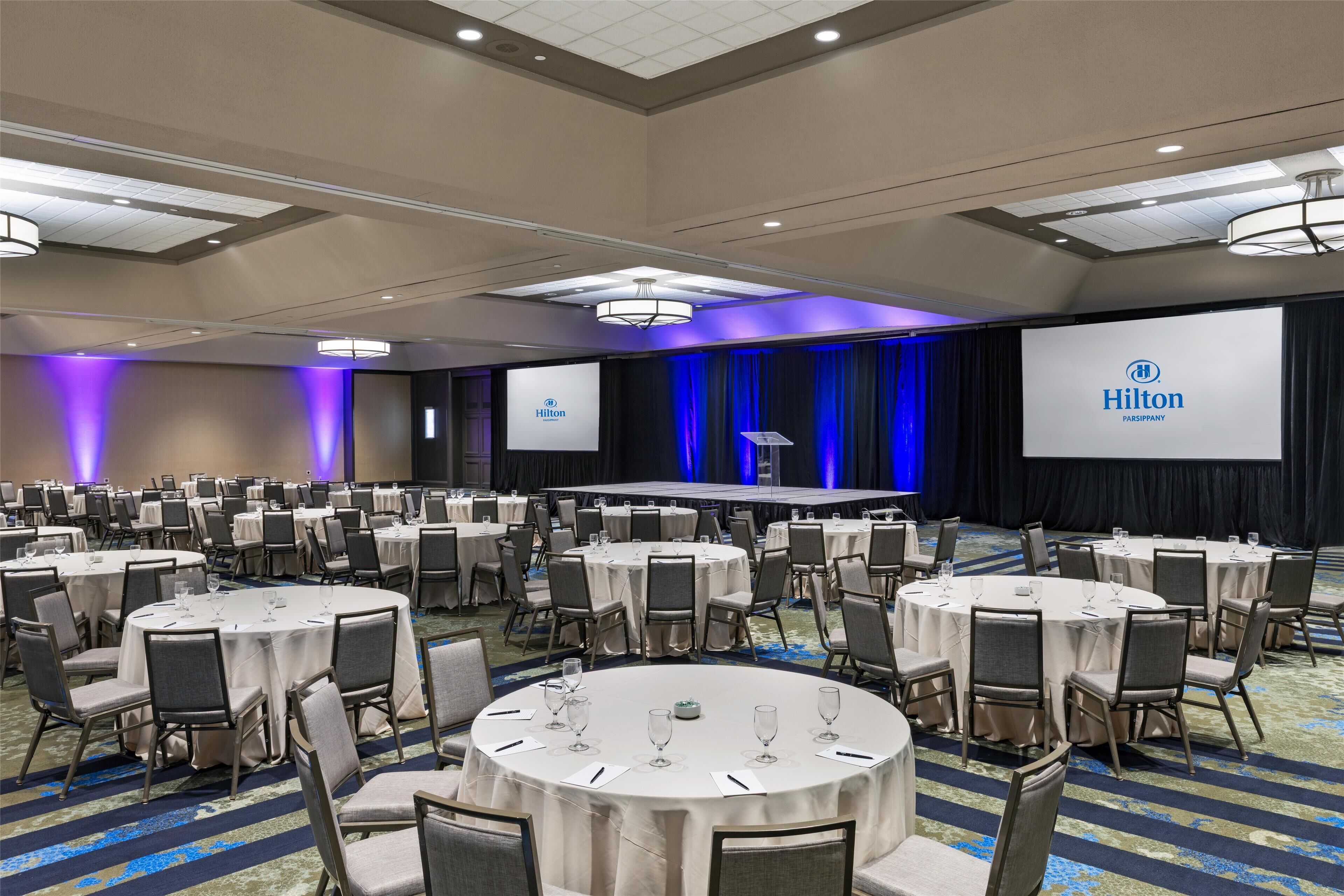 A large conference room with round tables and chairs, a stage with a podium, and a large screen displaying the Hilton logo.