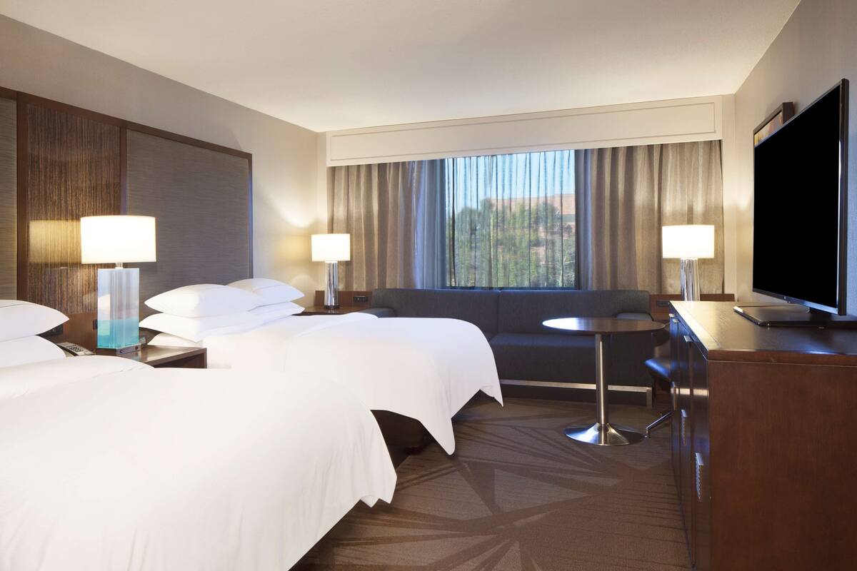 Quarto de hotel com duas camas, um sofá, uma escrivaninha, uma televisão e uma janela com cortinas.