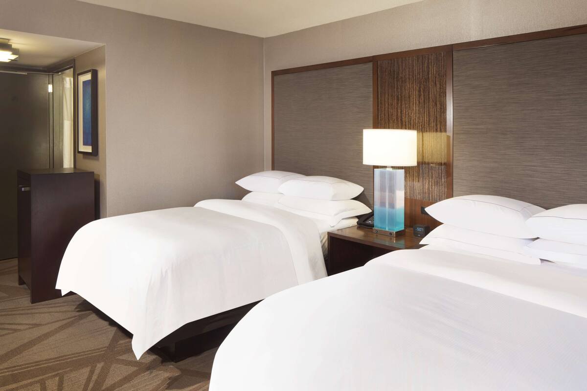 Quarto de hotel com duas camas, mesa de cabeceira com abajur, cômoda e um quadro na parede.