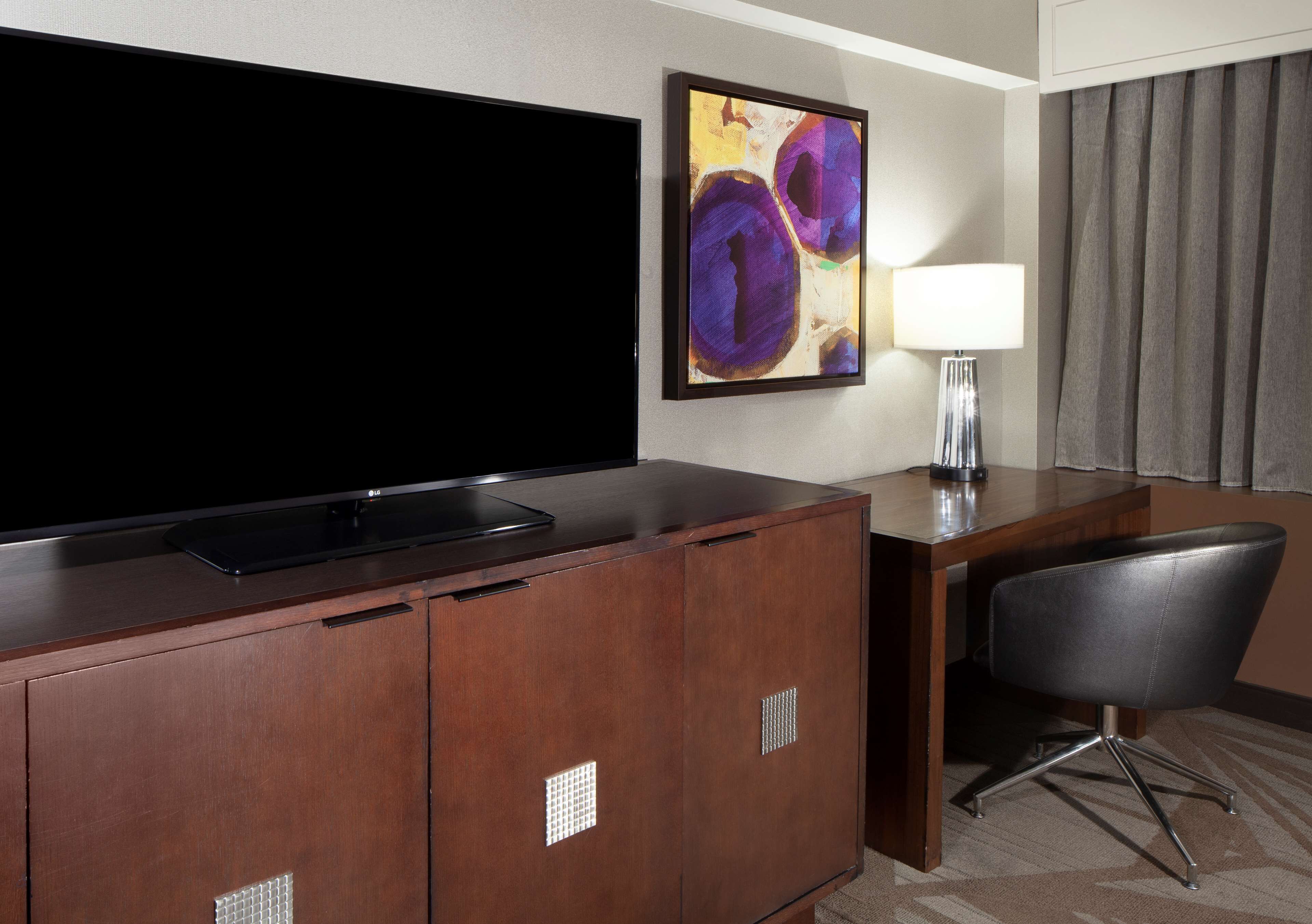 A hotel room with a large flat-screen TV, a wooden desk, a chair, a lamp, and a painting on the wall.