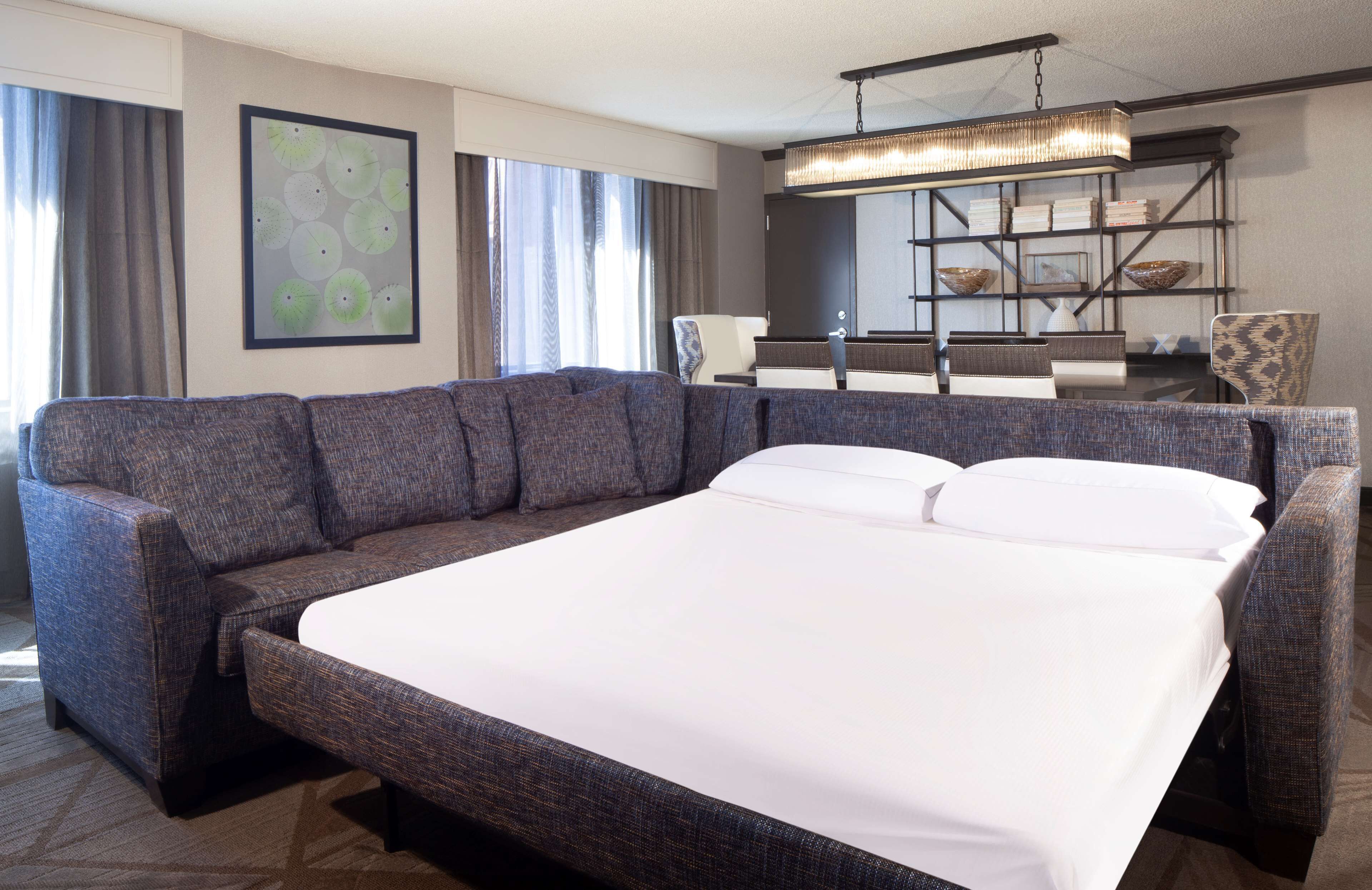 A hotel room with a large bed, a sofa, a dining table with chairs, a bookshelf, and a large window with curtains.