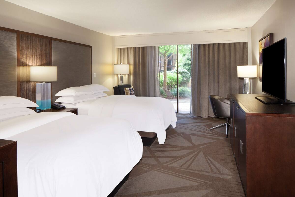 Quarto de hotel com duas camas, uma escrivaninha com televisão e vista para um jardim através de janelas amplas.