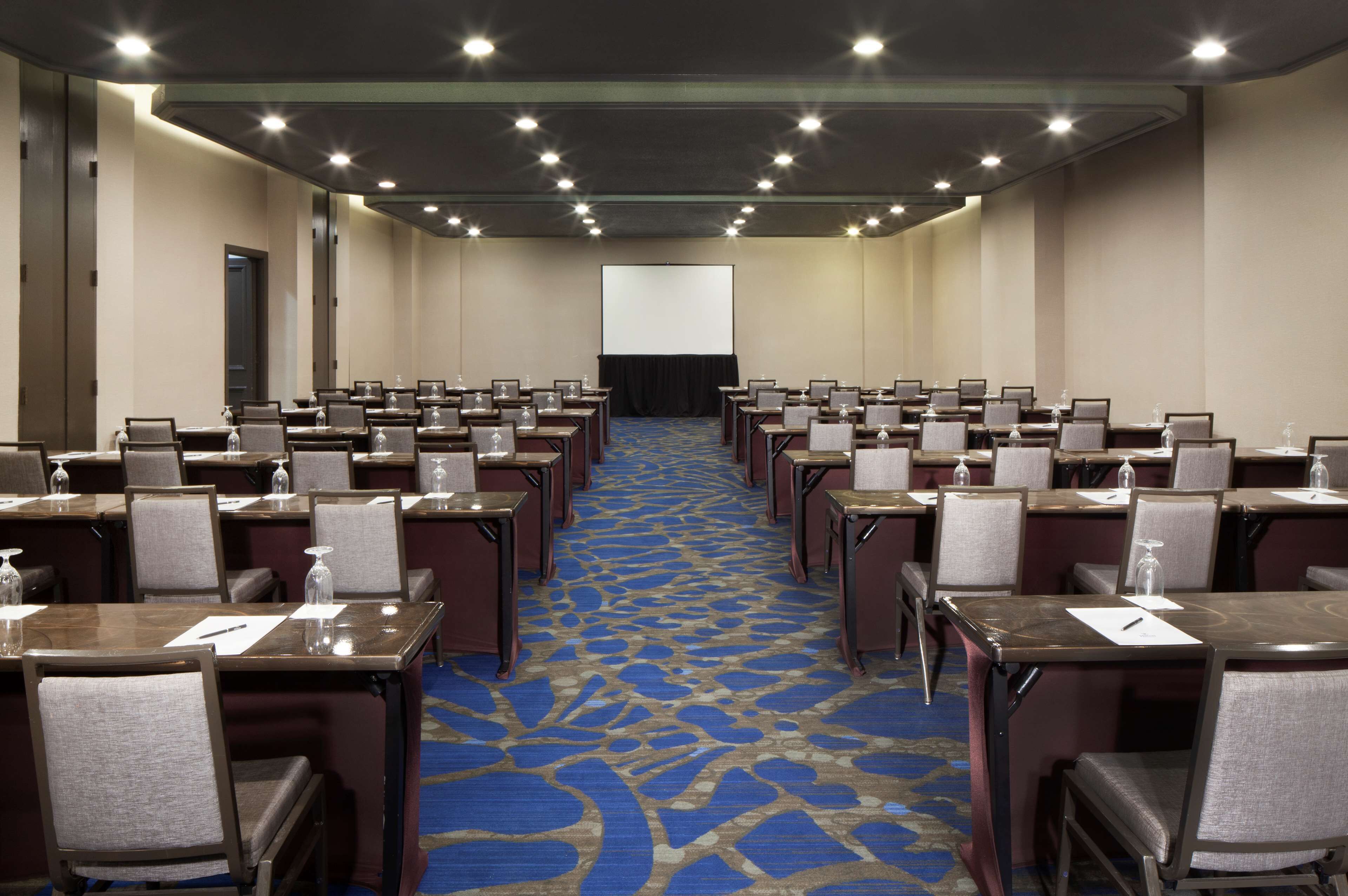 Un salón de conferencias con filas de mesas y sillas, una pantalla grande al frente y piso alfombrado.