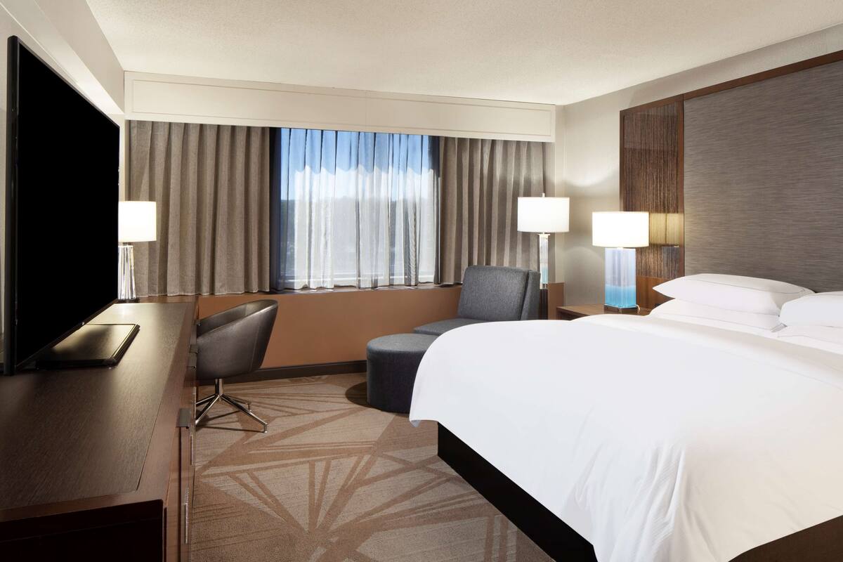 Quarto de hotel com uma cama grande, uma escrivaninha com cadeira, televisão e duas luminárias.