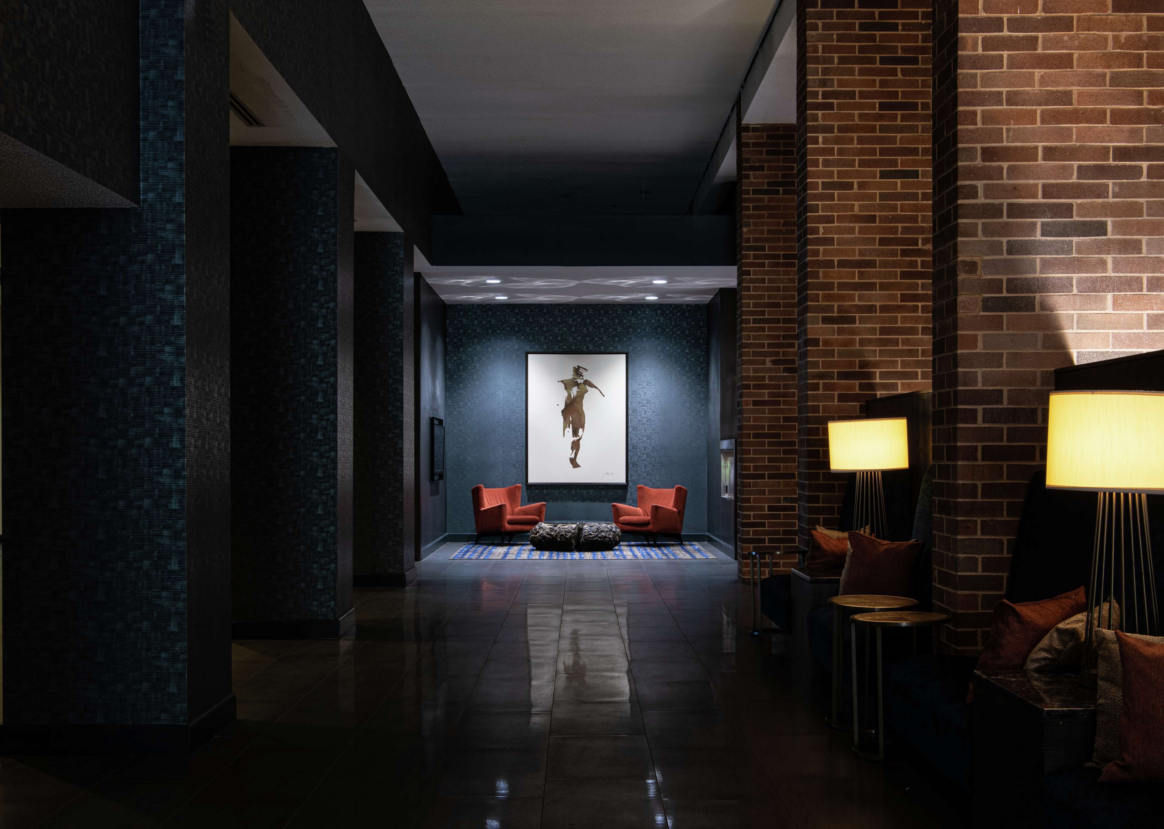 A modern lobby with a dark blue textured wall, a large abstract painting, two red armchairs, and a brick accent wall.