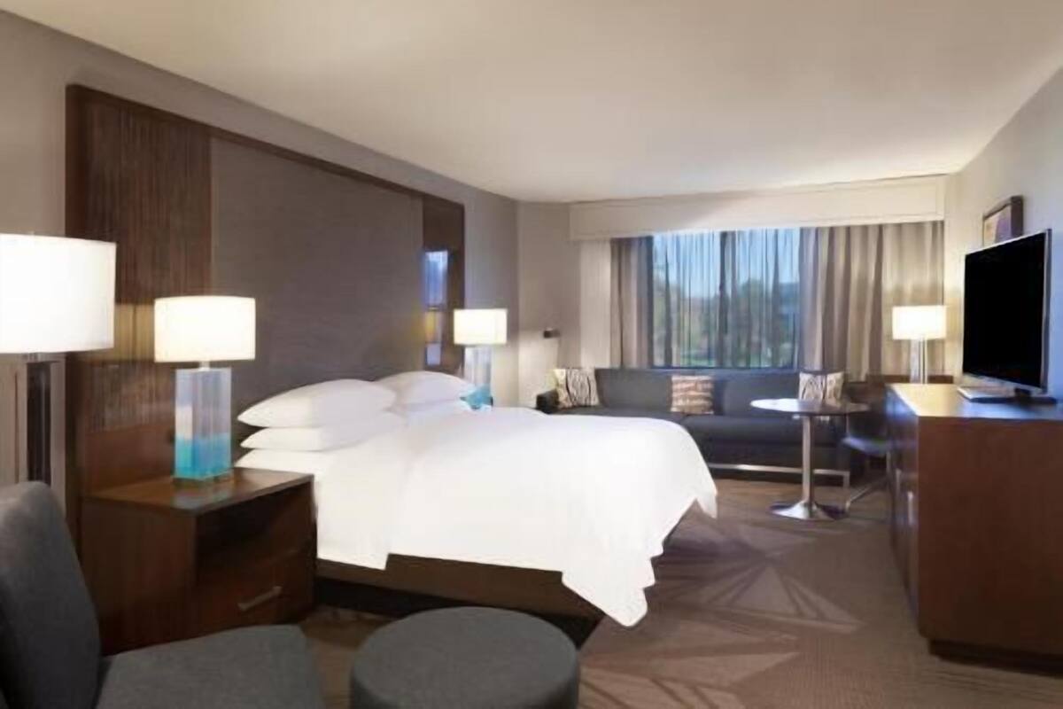 Quarto de hotel com cama, mesas de cabeceira, escrivaninha, cadeira, televisão e janela com cortinas.