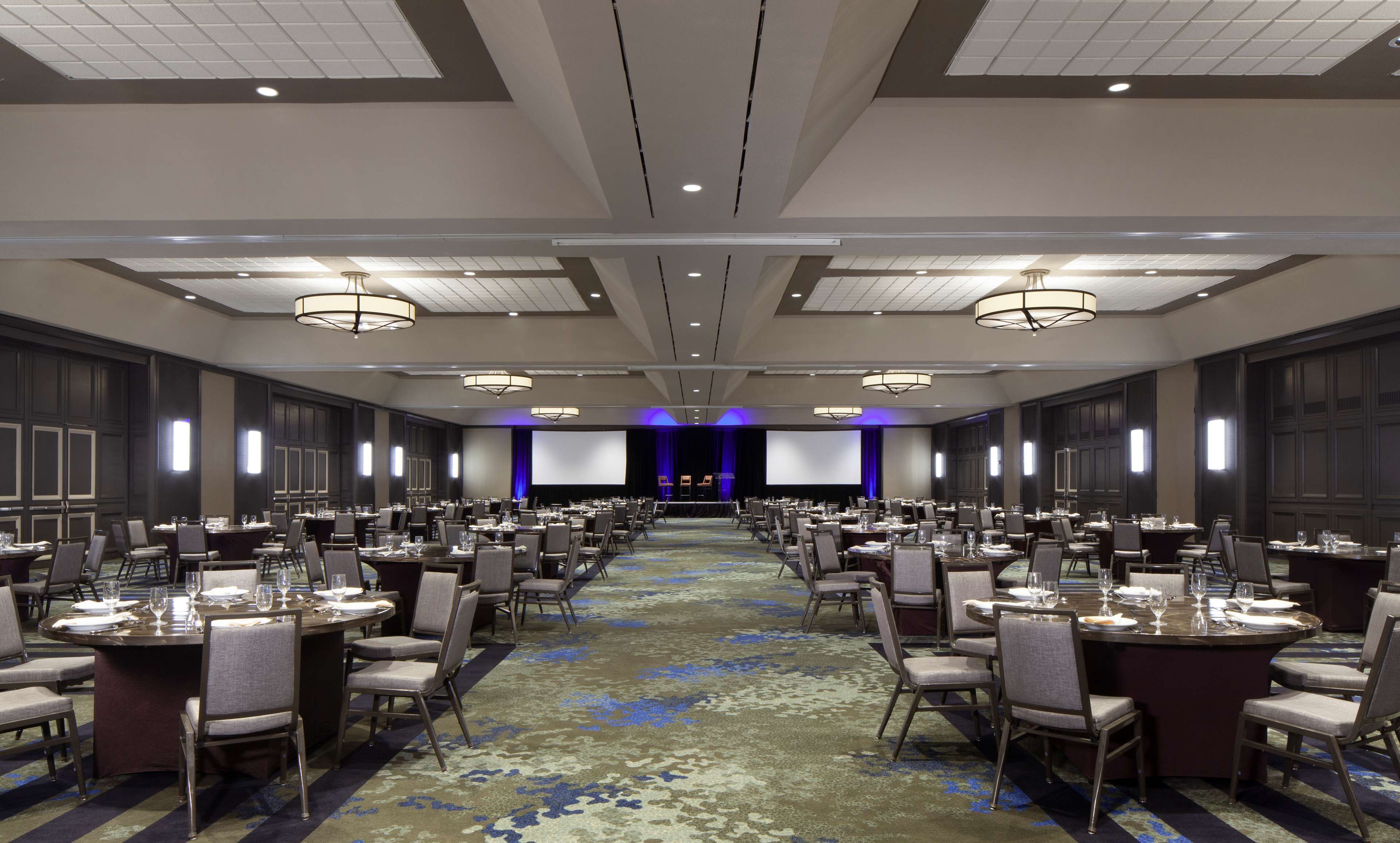 A large, well-lit conference room with multiple tables and chairs, a stage with a screen, and a carpeted floor.
