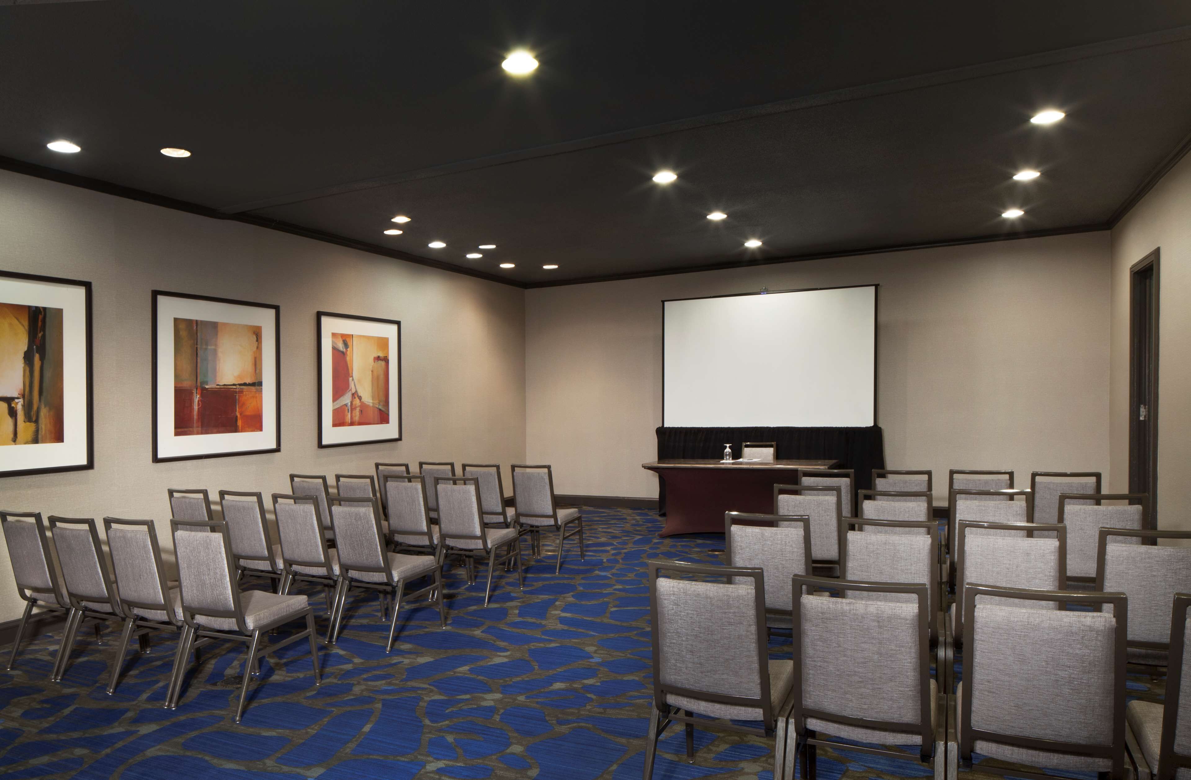 A conference room with rows of chairs, a podium, a projector screen, and framed artwork on the walls.
